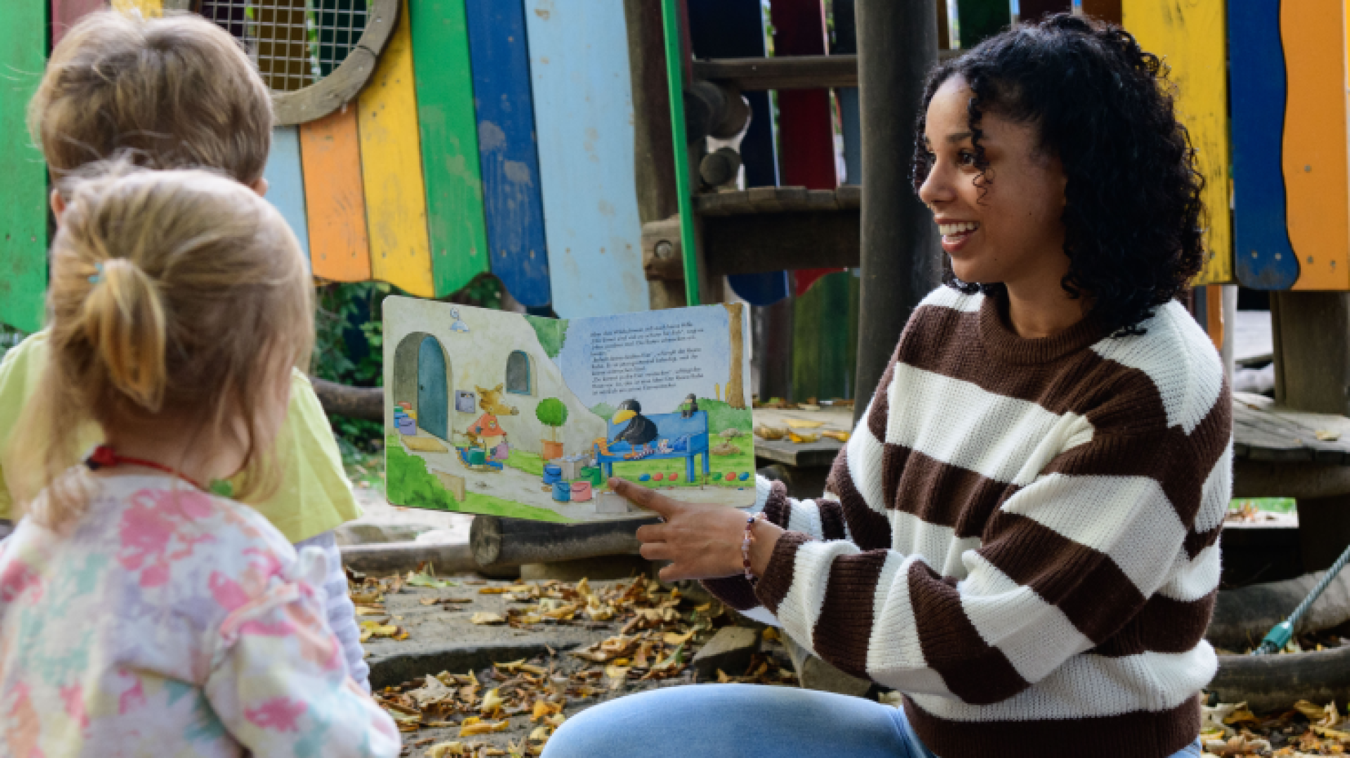 Eine Frau mit lockigem Haar liest zwei kleinen Kindern im Freien ein buntes Bilderbuch vor. Sie sitzen auf dem Boden neben einem Spielplatz mit Holzkonstruktionen und heruntergefallenen Blättern.